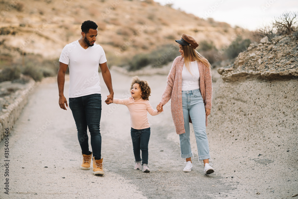 Happy diverse mother father and curly child wearing bright casual clothes strolling on nature holding hands at daytime Happy diverse mother father and curly child wearing bright casual clothes strolling on nature holding hands at daytime