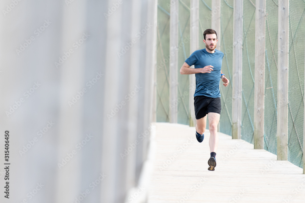 Highly motivated bearded male athlete in blue t shirt and shorts running outdoors under cover looking at camera Highly motivated bearded male athlete in blue t shirt and shorts running outdoors under cover looking at camera