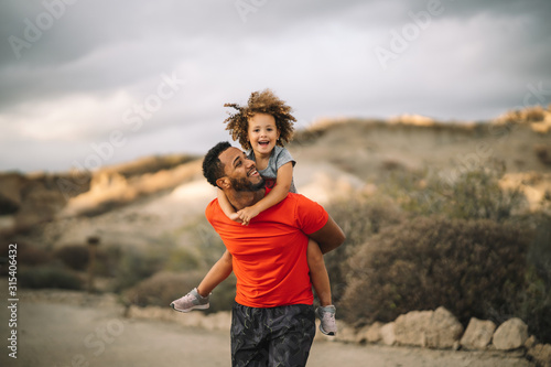 Smiling African American bearded man dressed in sportive clothes carrying cheerful curly active toddler while walking on nature photo