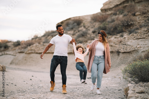 Happy diverse mother father and curly child wearing bright casual clothes strolling on nature holding hands at daytime photo