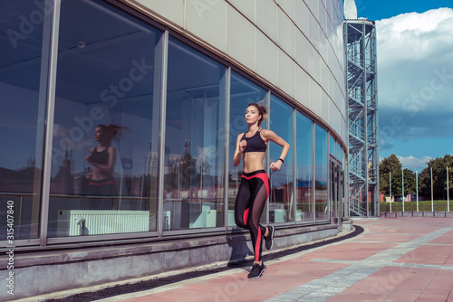 Beautiful girl runs in summer in city, jogging in early morning and afternoon, fitness training in fresh air. Free space for copy text. Sportswear, tanned figure. Healthy lifestyle of a woman. © byswat