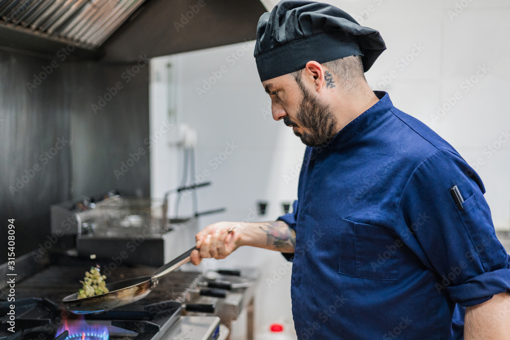 Side view of male cook in black hat shaking frying pan while cooking vegetables for dish on gas stone in restaurant kitchen Side view of male cook in black hat shaking frying pan while cooking vegetables for dish on gas stone in restaurant kitchen