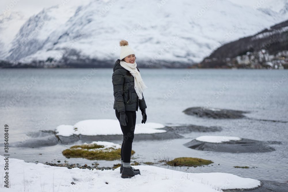 Profile view of young cheerful asian woman in warm down jacket and hat standing and looking at camera on beach against white and brown snowy hills and tranquil cool water of Kaldfjorden lake in Norway Profile view of young cheerful asian woman in warm down jacket and hat standing and looking at camera on beach against white and brown snowy hills and tranquil cool water of Kaldfjorden lake in Norway