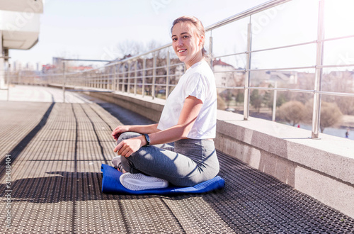 Young athletic woman in a white T-shirt and gray leggings sits in a lotus position in the fresh air in sunny weather on the terrace. Smile and look at camera