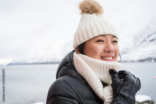 Happy female with amazing eyes in black jacket and white warm hat and scarf looking away and smiling against white blurred background in Norway photo