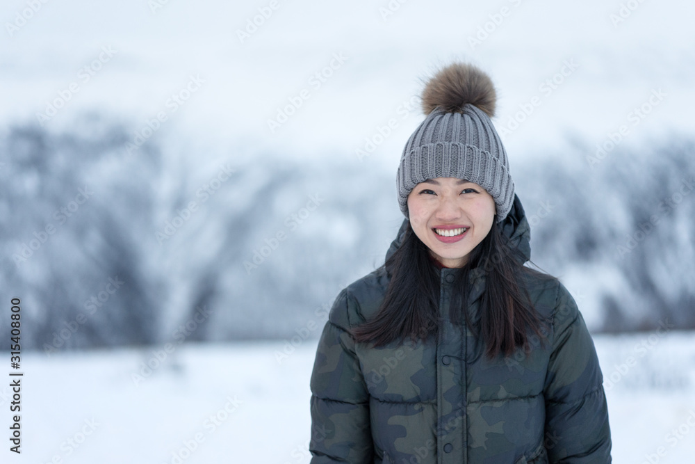 Magnificent happy female with amazing eyes in dark spotted down jacket and gray warm hat looking at camera and smiling against blurred scenery of snowy park in daytime Magnificent happy female with amazing eyes in dark spotted down jacket and gray warm hat looking at camera and smiling against blurred scenery of snowy park in daytime