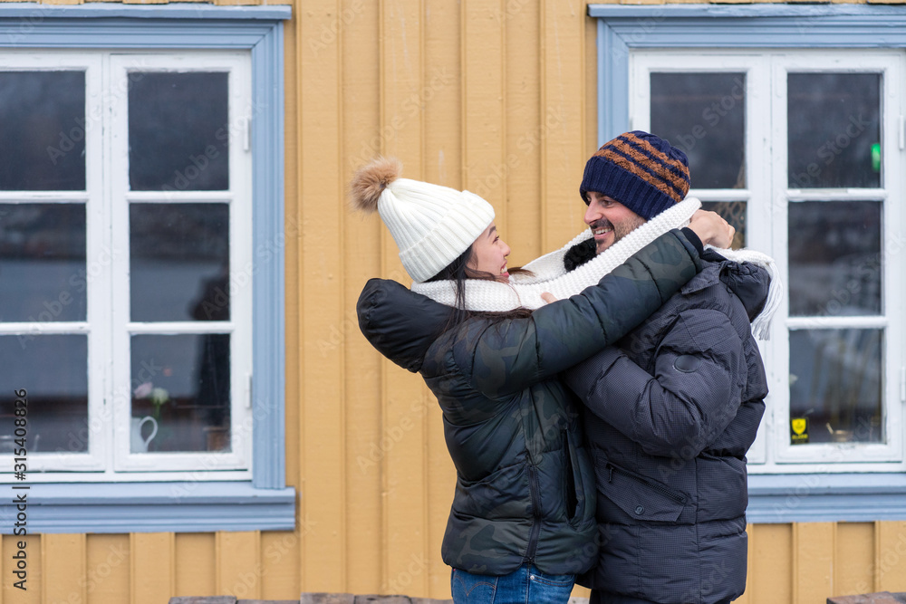 Side view of multiethnic cheerful couple in warm clothing standing and hugging with rural house on background at Tromso in Norway Side view of multiethnic cheerful couple in warm clothing standing and hugging with rural house on background at Tromso in Norway