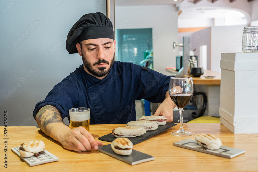 Serious professional male cook in uniform and hat arranging tray with gourmet sandwiches while standing at counter with drinks and snacks ready for serving Serious professional male cook in uniform and hat arranging tray with gourmet sandwiches while standing at counter with drinks and snacks ready for serving