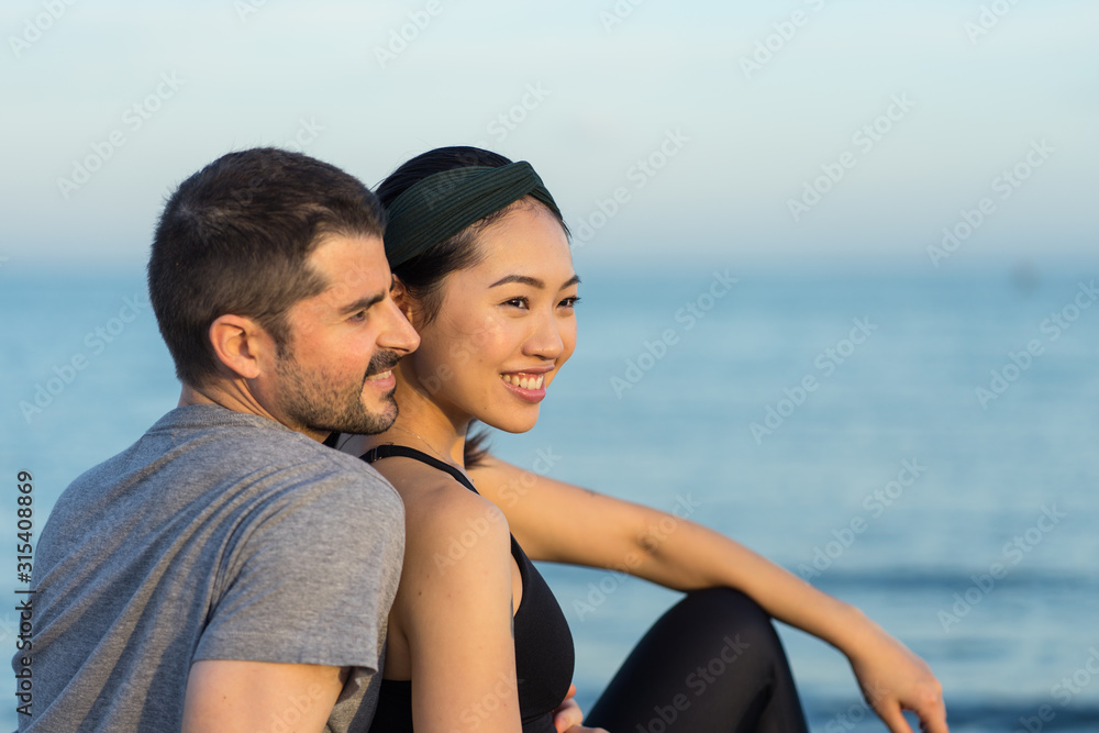Side view of cheerful young multiracial couple in sportswear sitting on sandy beach while resting after training and enjoying time together