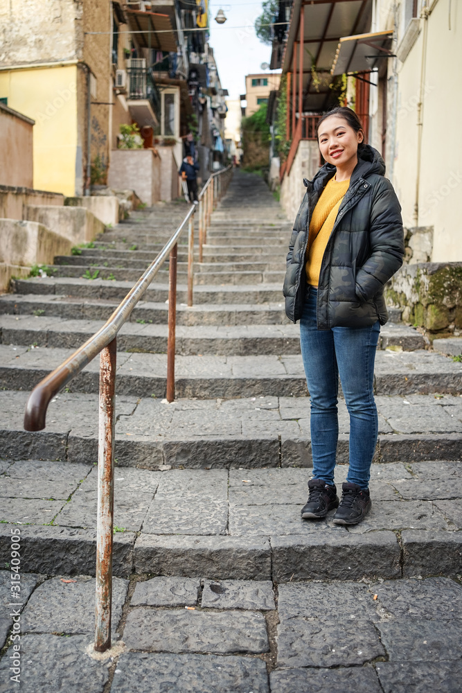 Full body Asian female with hands in pockets looking at camera while standing on stairs outside weathered buildings on city street Full body Asian female with hands in pockets looking at camera while standing on stairs outside weathered buildings on city street