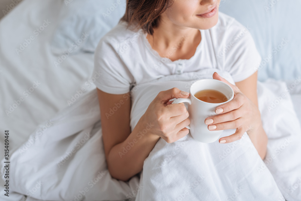 cropped view of dreamy woman holding cup with tea in bed cropped view of dreamy woman holding cup with tea in bed