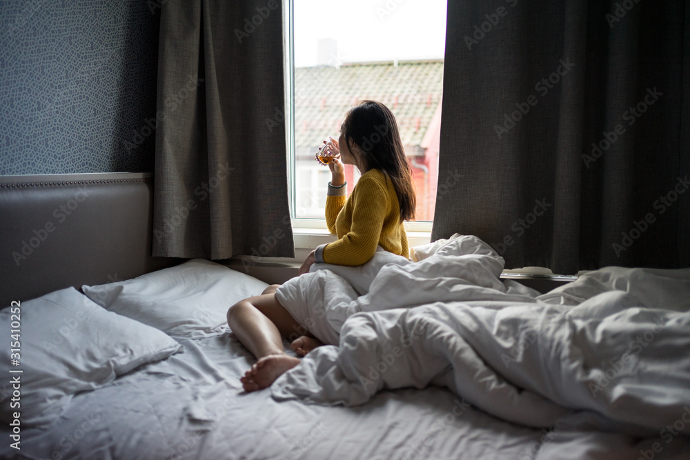 Side view of black haired faceless female in yellow sweater sitting on bed while drinking tea and looking at window on lazy morning at Tromso in Norway Side view of black haired faceless female in yellow sweater sitting on bed while drinking tea and looking at window on lazy morning at Tromso in Norway