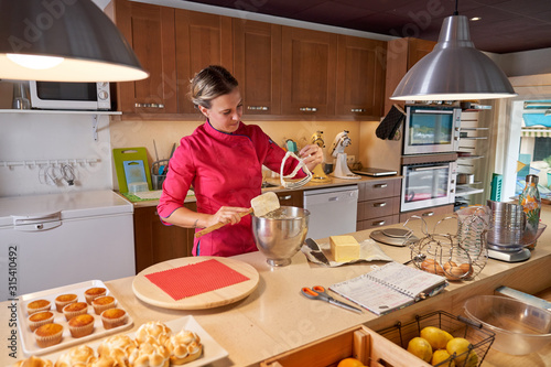 Attentive adult cook in stylish vivid pink uniform cleaning with spatula whisk after beating cream in metallic bowl while working at table beside wooden cake stand and other useful kitchen stuff at home