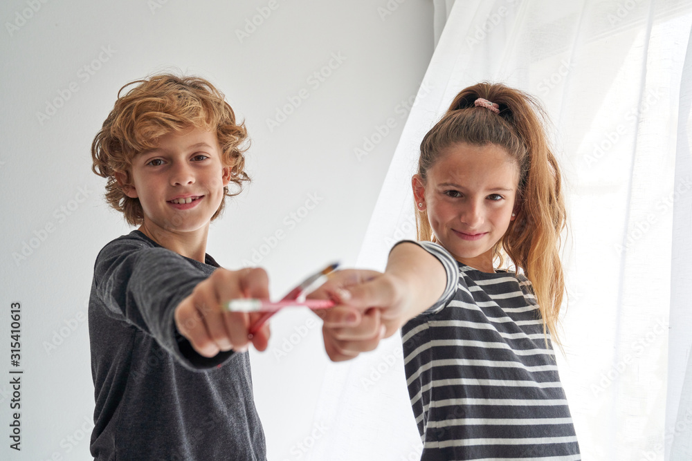 Delighted boy and girl in casual outfit looking at each other and crossing paintbrushes while standing on chairs near table with watercolor and playing at home Delighted boy and girl in casual outfit looking at each other and crossing paintbrushes while standing on chairs near table with watercolor and playing at home