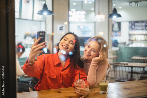 Two young woman friends with cups of fresh coffee smiling and posing for selfie while sitting at table in cozy restaurant having a beverage photo