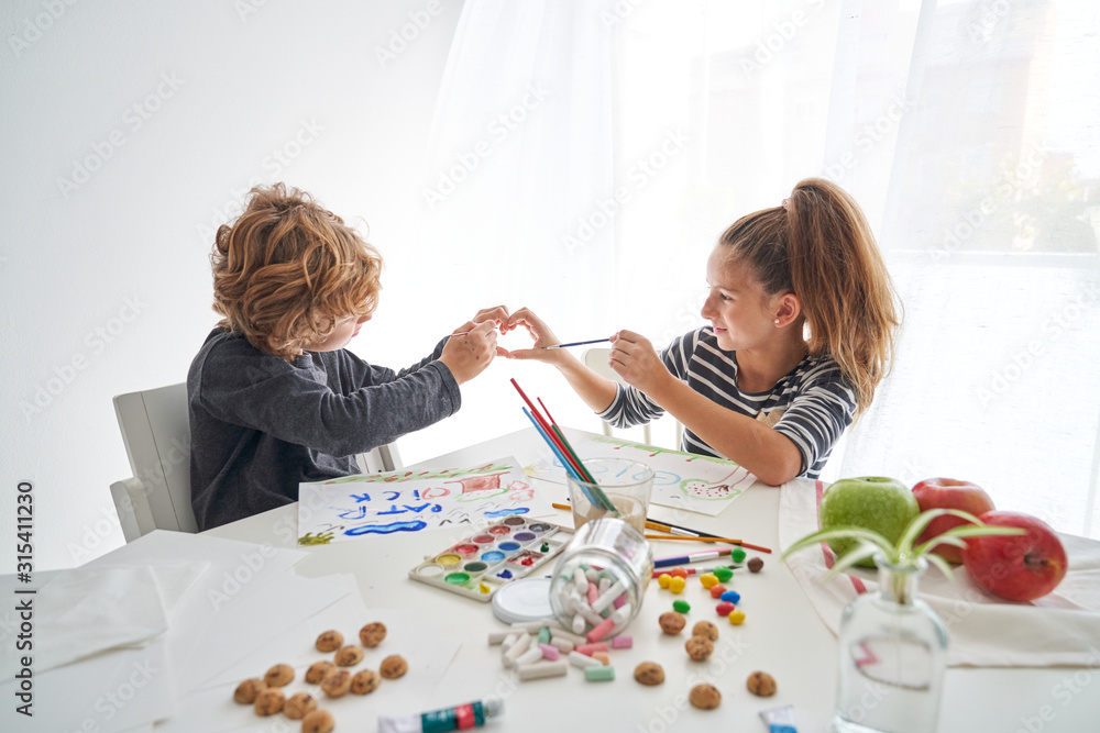 Little girl and boy smiling and showing heart gesture while sitting at table and painting at home together Little girl and boy smiling and showing heart gesture while sitting at table and painting at home together