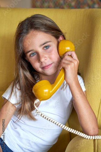 Little girl in white t shirt and jeans sitting on yellow comfortable armchair and talking on fusty telephone and looking at camera