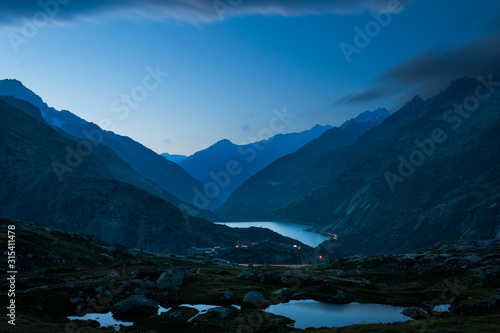 Mysterious dark blue mountain range and river between slopes with burning lights along road in Switzerland