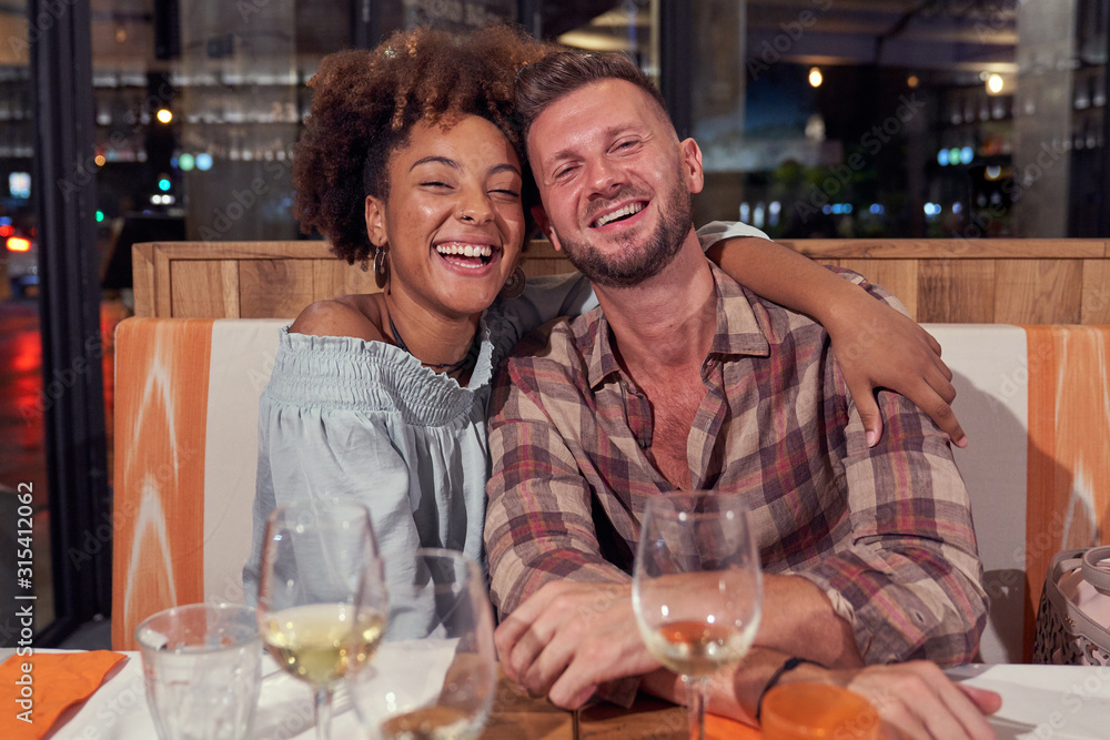 Multiethnic couple in casual clothing smiling while sitting at festive table in am embrace and looking at camera at restaurant Multiethnic couple in casual clothing smiling while sitting at festive table in am embrace and looking at camera at restaurant