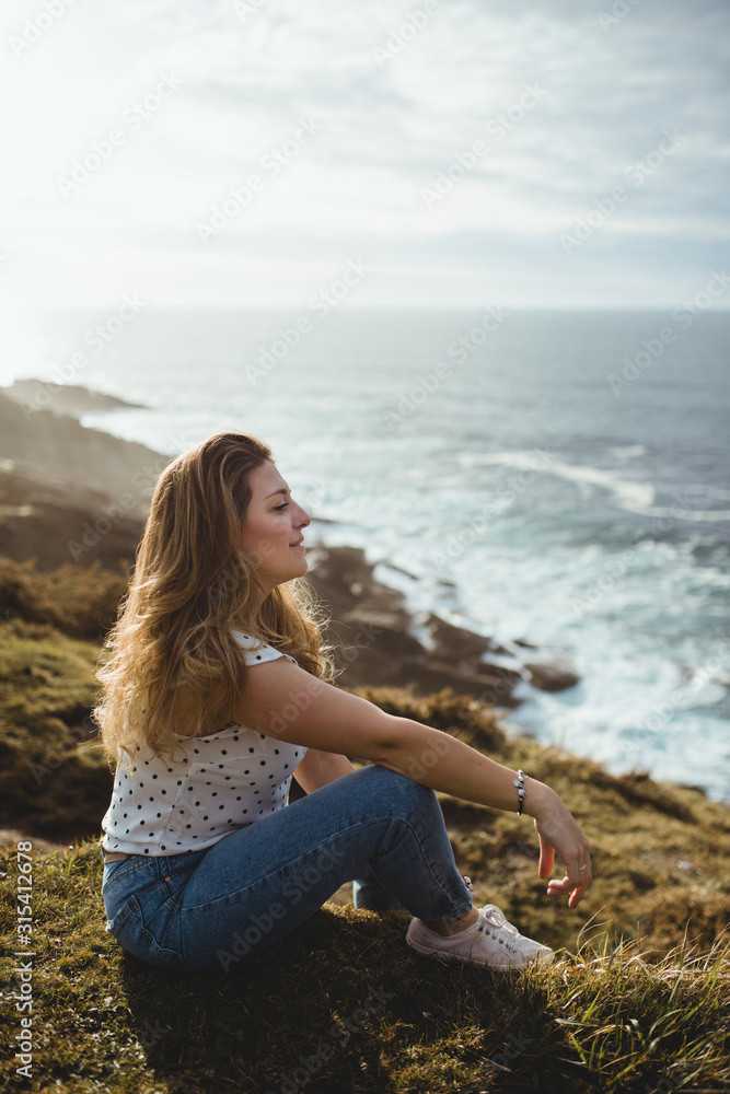 Side view of young female in casual clothes relaxing and dreaming while sitting on grass and looking at sea waves in sunny day with clouds on sky Side view of young female in casual clothes relaxing and dreaming while sitting on grass and looking at sea waves in sunny day with clouds on sky