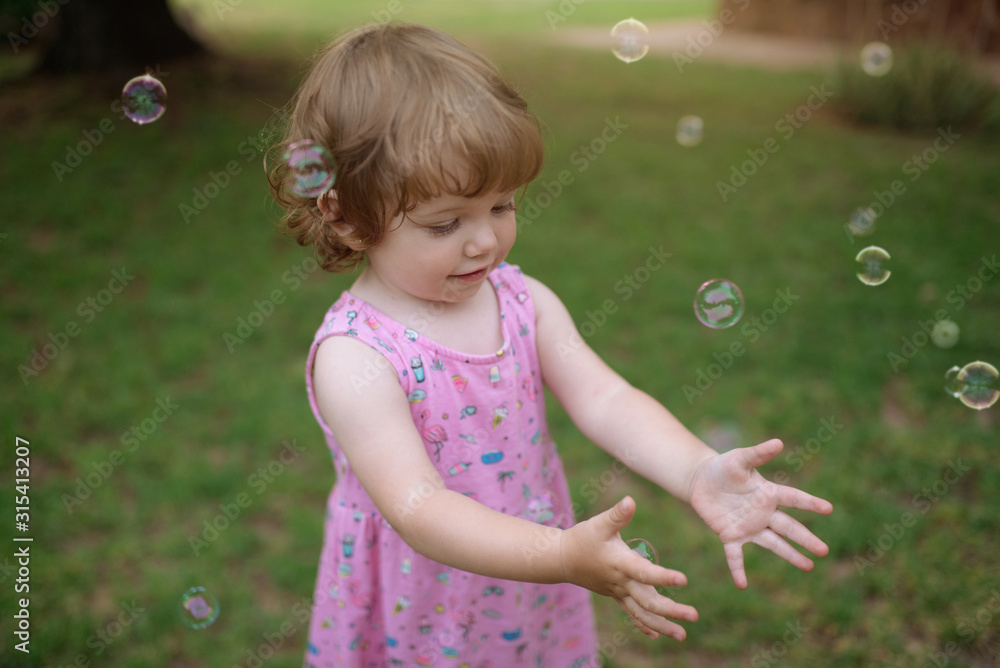 From above adorable child in pink dress laughing and capturing rainbow soap bubbles on green meadow in park From above adorable child in pink dress laughing and capturing rainbow soap bubbles on green meadow in park
