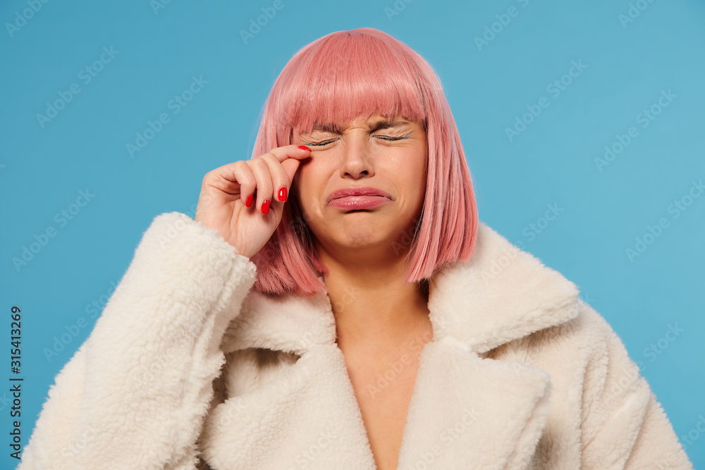 Unhappy young pretty pink haired female with bob haircut frowning her face while going to cry, keeping eyes closed and raisind hand to her face, standing against blue background Unhappy young pretty pink haired female with bob haircut frowning her face while going to cry, keeping eyes closed and raisind hand to her face, standing against blue background