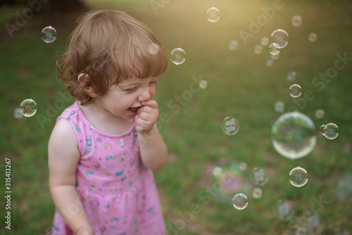 From above adorable child in pink dress laughing and capturing rainbow soap bubbles on green meadow in park photo