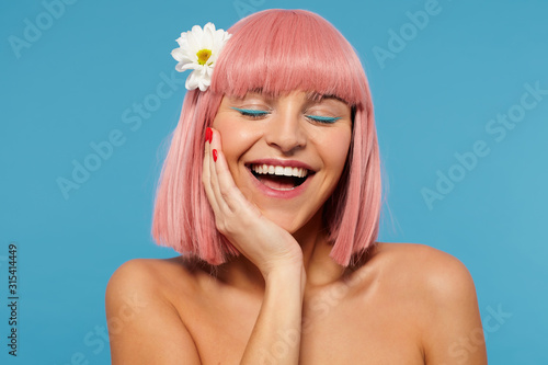 Indoor photo of pleasant looking happy young female with short pink hair keeping her eyes closed while leaning her head on raised hand, smiling cherfully over blue background photo