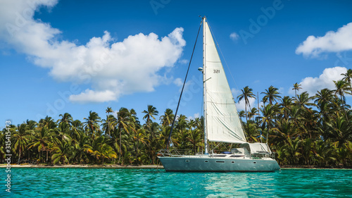 A grey-white Sailing Yacht anchored in Turquoise Water in front of the paradisiacal San Blas Islands in Panama with green Palm Trees and a perfect Blue Sky in the background. © Stefan