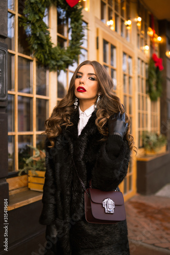 Beautiful young caucasian woman wearing natural dark fur coat posing for a portrait near Christmas showcase. Christmas decor. Fashion. Style. Winter clothes made of natural fur. Vertical