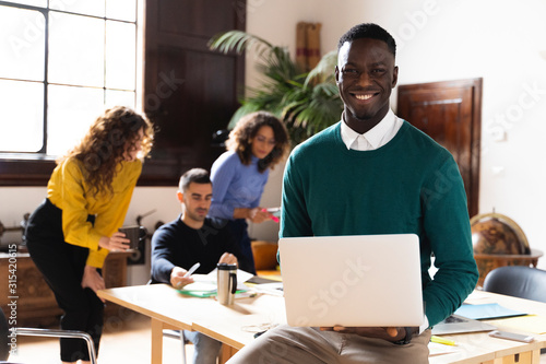 Portrait of a smiling man in office using laptop with colleagues in background photo