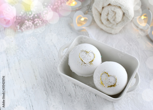 Romantic spa set on white background. Close up bath bombs in ceramic bowl with hearts, towel on wooden desk. Bokeh effect. Copyspace.