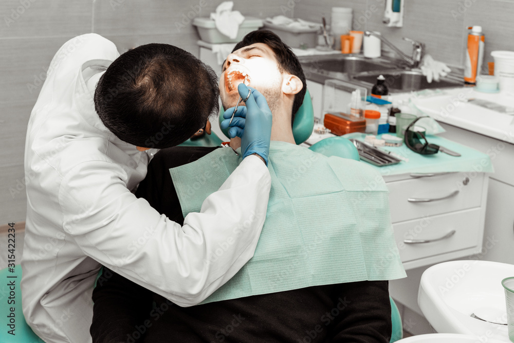 A young male dentist doctor treats a patient. Medical manipulations in dentistry, surgery. Professional uniform and equipment of a dentist. Healthcare Equipping a doctor’s workplace. Dentistry A young male dentist doctor treats a patient. Medical manipulations in dentistry, surgery. Professional uniform and equipment of a dentist. Healthcare Equipping a doctor’s workplace. Dentistry