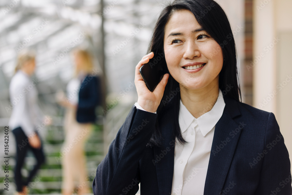 Portrait of a smiling businesswoman on the phone in office with colleagues in background Portrait of a smiling businesswoman on the phone in office with colleagues in background