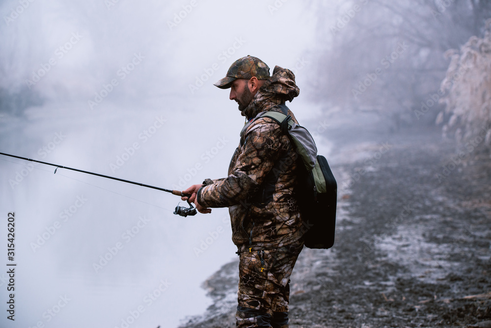Fisher man fishing with spinning rod on a river bank at misty foggy winter, spin fishing, prey fishing Fisher man fishing with spinning rod on a river bank at misty foggy winter, spin fishing, prey fishing