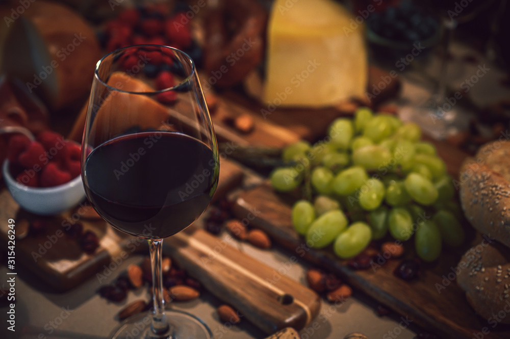 Glass of red wine accompanied by a variety of gourmet ingredients such as a variety of cheeses, cold meats, wine bottles, rustic bread and fruit on a rustic background
