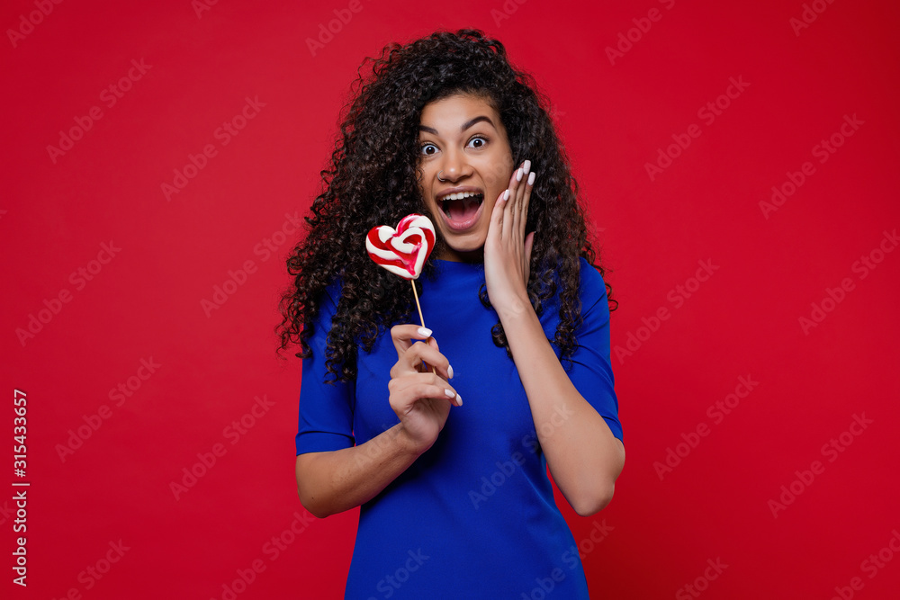 black woman smiling with heart shaped lollypop candy isolated on red background black woman smiling with heart shaped lollypop candy isolated on red background