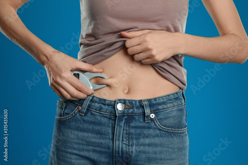 Young woman measuring body fat with caliper on blue background, closeup