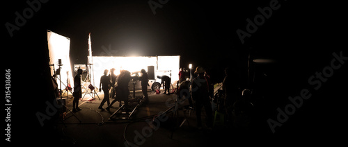 Silhouette images of video production behind the scene or b-roll or making of film shooting and movie crew team working in back of camera and lighting soft box equipment set at outdoor in the night