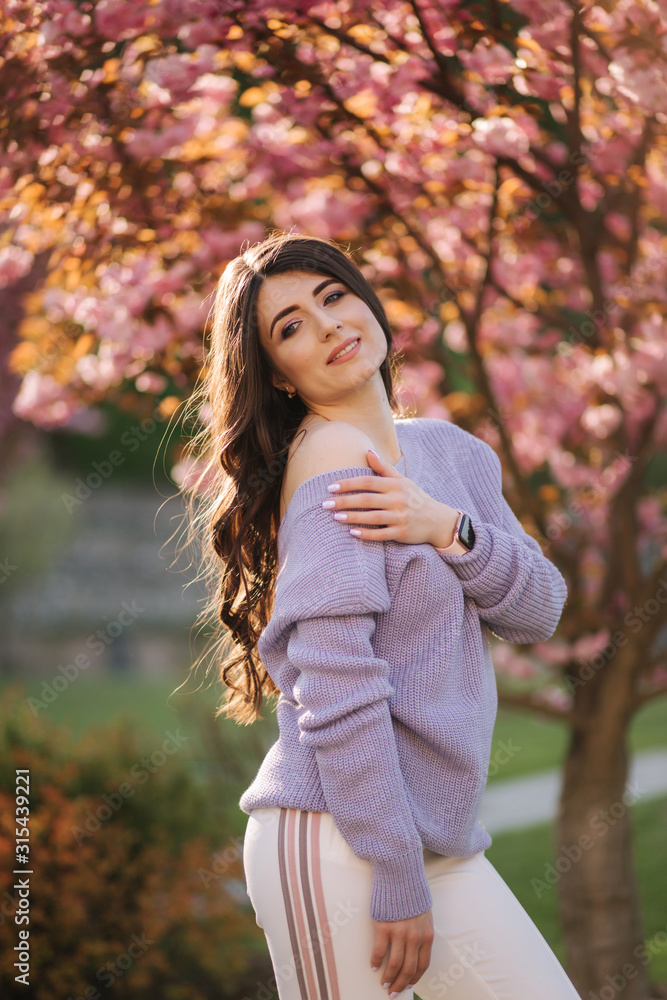 Attractive young woman in a purple sweater stand in front of sakura tree in the park Attractive young woman in a purple sweater stand in front of sakura tree in the park