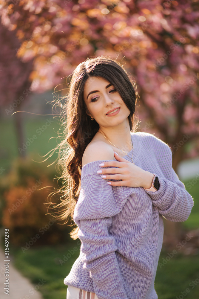 Attractive young woman in a purple sweater stand in front of sakura tree in the park Attractive young woman in a purple sweater stand in front of sakura tree in the park