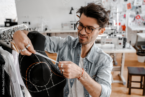 Attractive professional serious stylish tailor with glasses in a blue shirt working next to a wooden table with threads, an apron and scissors in an amazing modern studio and mannequin.