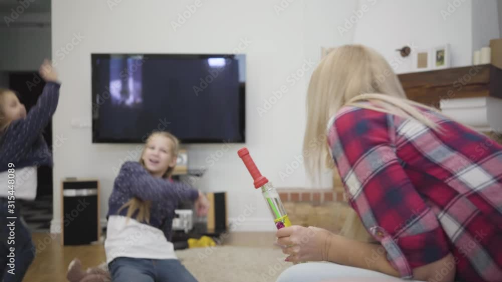 Back view of blond Caucasian woman blowing soap bubbles for daughters. Cheerful girls laughing and trying to catch bubbles. Happy family resting at home. Happiness, leisure, unity.