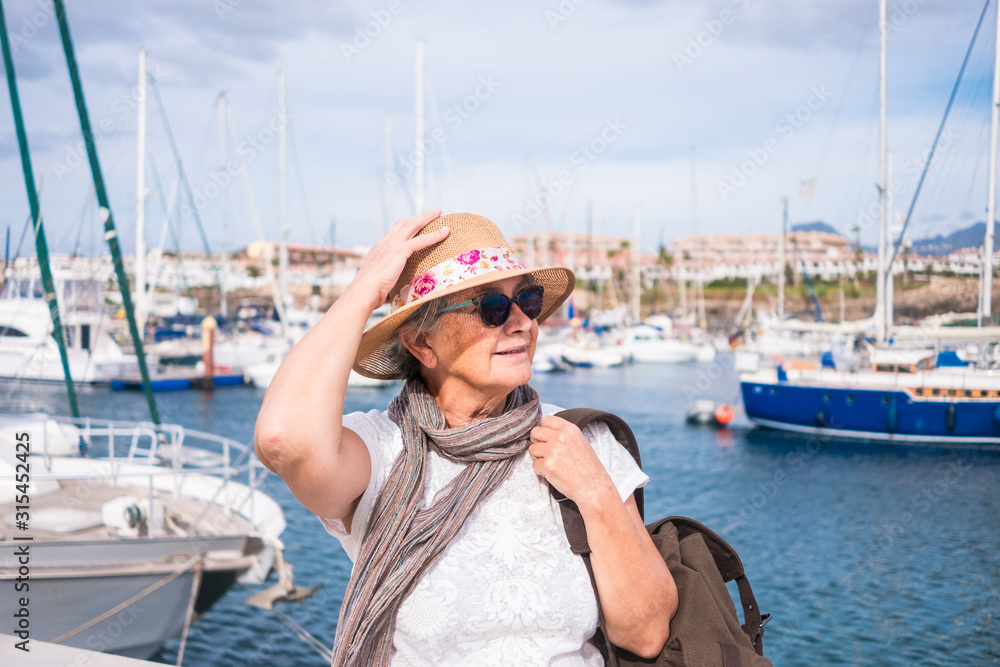 Portrait of an elderly woman with gray hairs and hat sitting at the port. A small port with yachts and sailboats. Cloudy and windy day