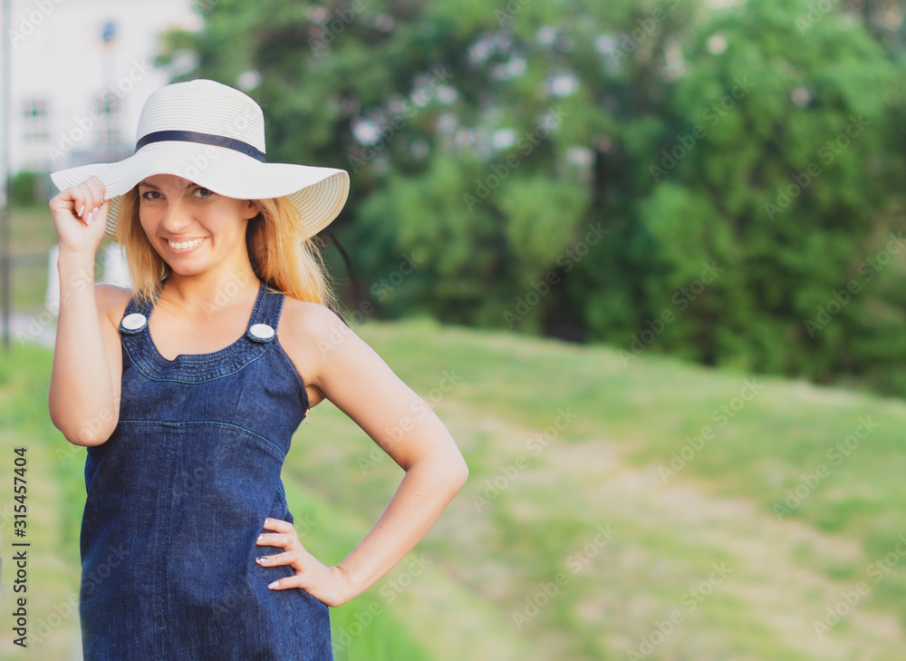 Beautiful young fashionable girl with a happy smile in a sun hat and a denim sundress on a green Park background on a summer day. Copy space.