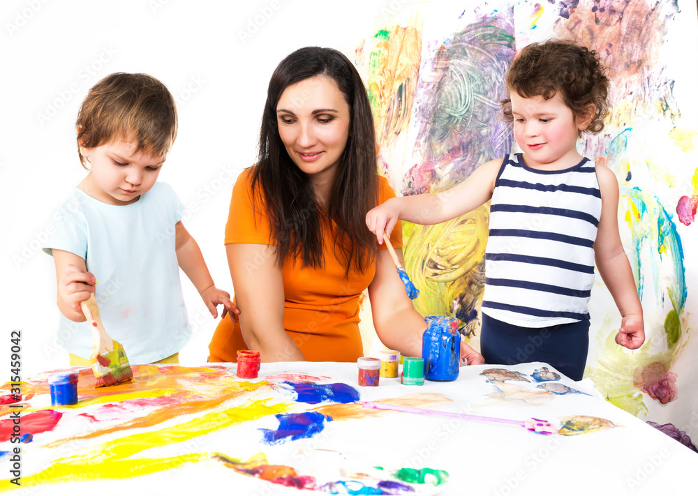 Woman with two babies painting with paints and brushes while sitting at childrens table, colorful background Woman with two babies painting with paints and brushes while sitting at childrens table, colorful background
