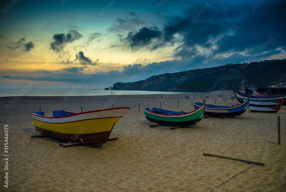 Fototapeta premium Portugal, twilight over Nazare beach , colored wooden boats, Traditional Portuguese fishing boats in Nazare on the coast, blue clouds on the sky, white sand of beaches of Portugal