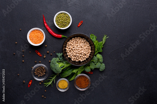 A variety of legumes chickpeas, lentils, mung bean in white bowls with spinach leaves, broccoli and spices on a black background. Background menu food.