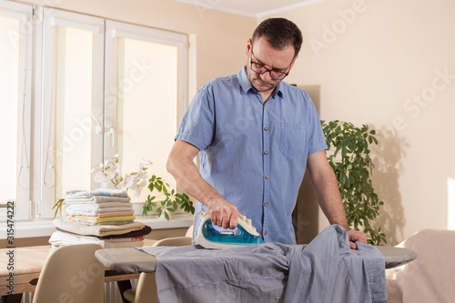 Mężczyzna w okularach ubrany w koszulę z krótkim rękawem podczas prasowania żelazkiem koszuli na desce do prasowania.