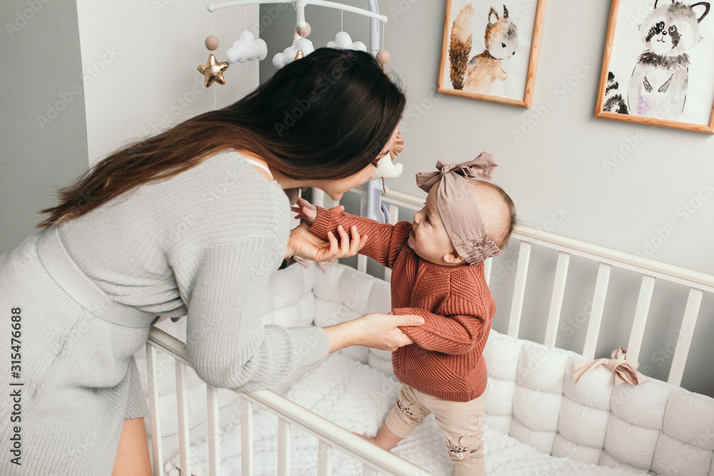 Beautiful baby girl in bed with her mom, light stylish interior. trying to stand. Concept of matherhood.